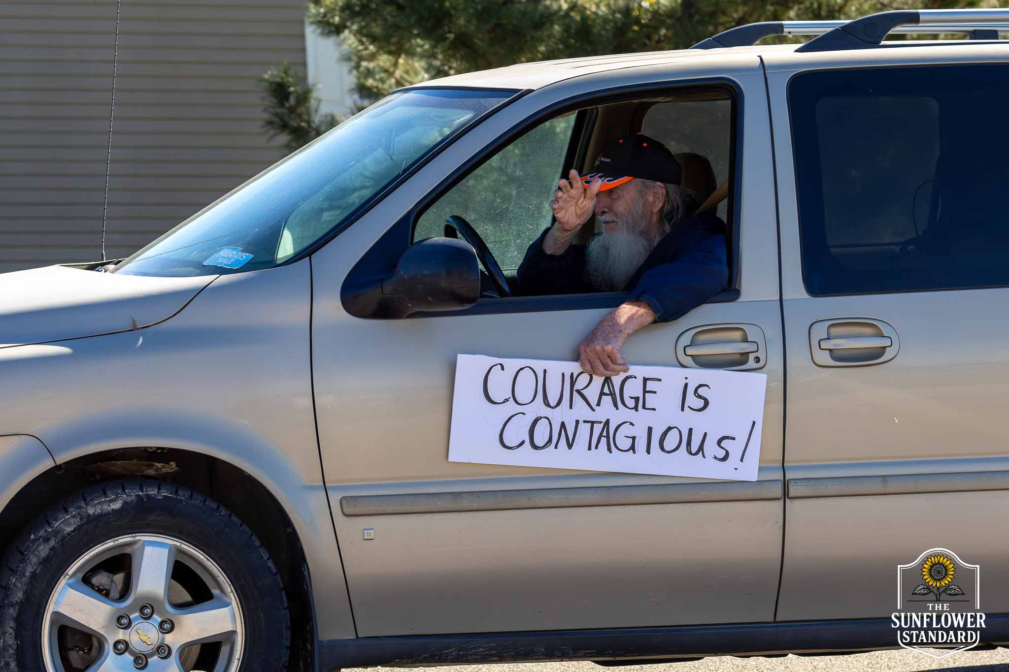 Ken Poland waves from a vehicle displaying a sign that reads Courage is contagious